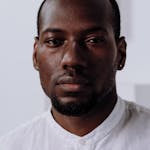 Headshot of a serious black man in a white shirt, indoors with a neutral background.